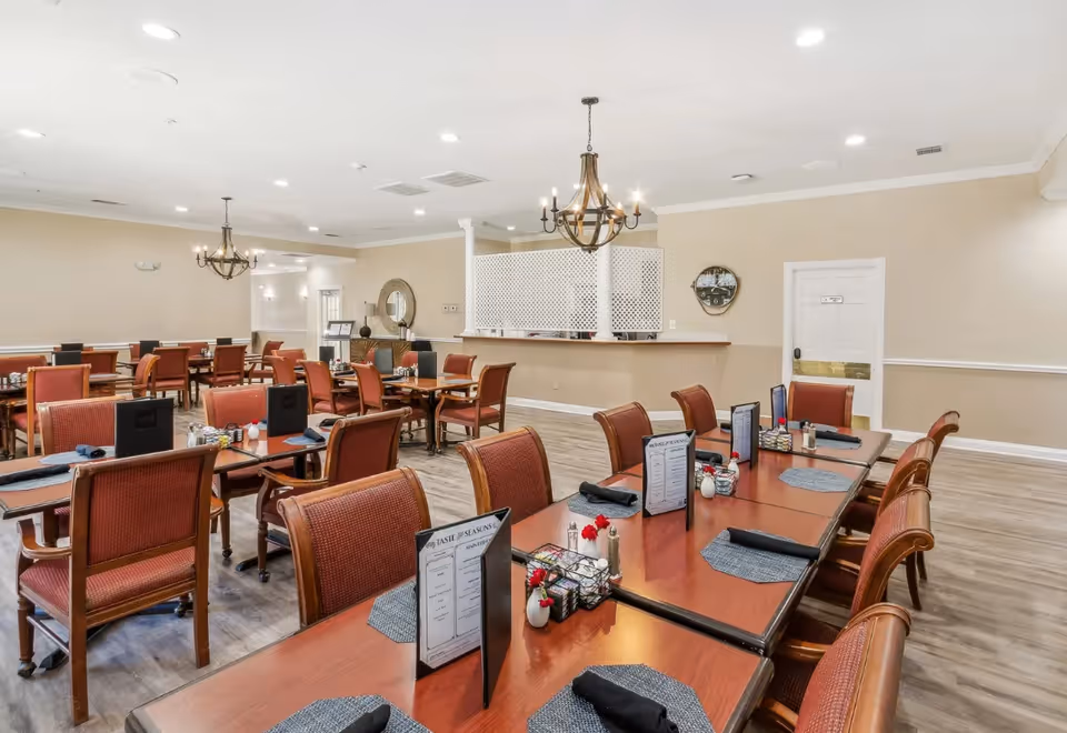 Spacious dining room with multiple wooden tables and red upholstered chairs set for meals under chandeliers.