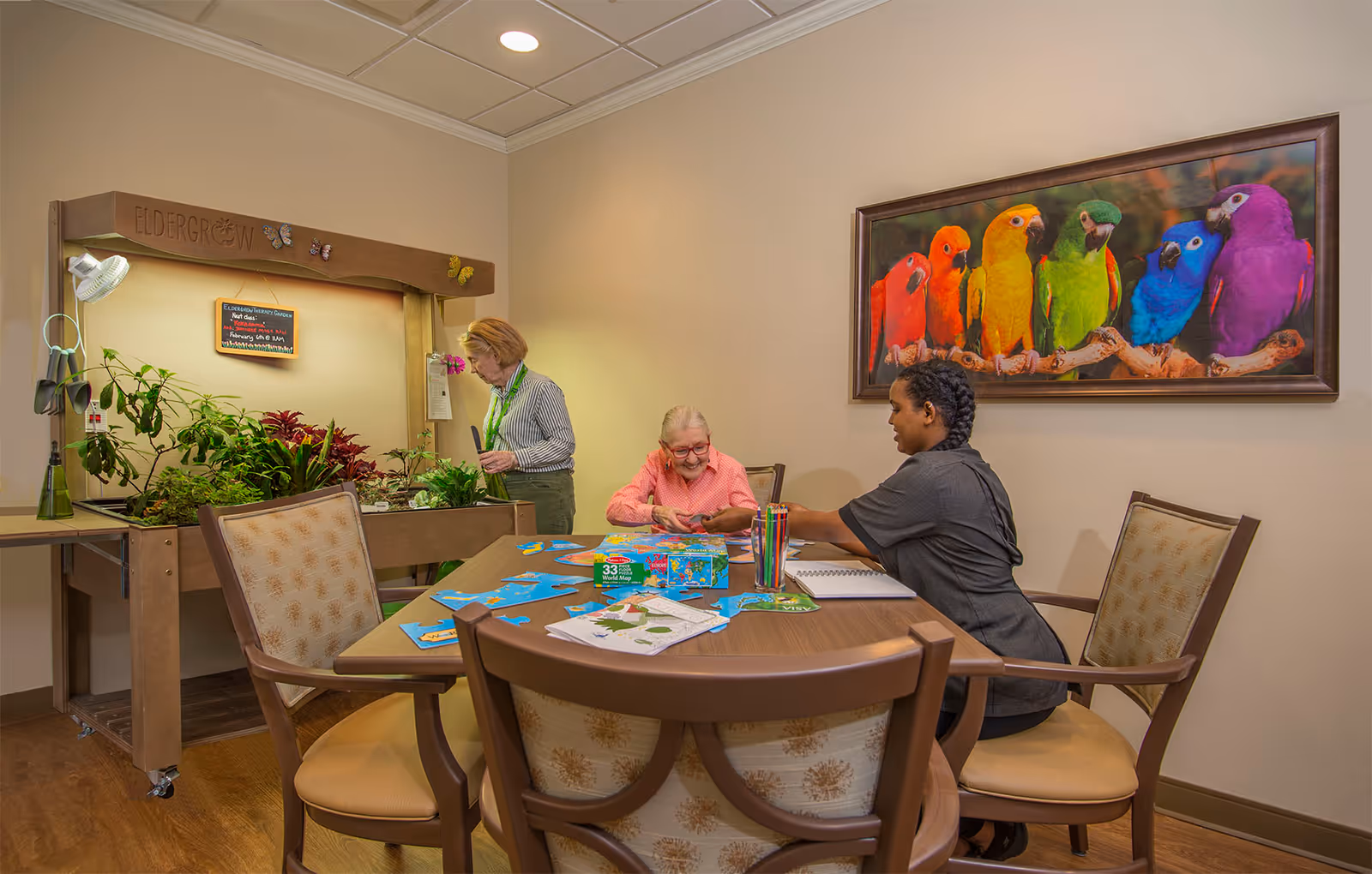 A senior woman and a caregiver sitting at a table working on a puzzle together in a well-lit room. Another senior woman is tending to plants in a small indoor garden labeled 'Eldergrow'. A colorful framed picture of six parrots is hanging on the wall behind them.