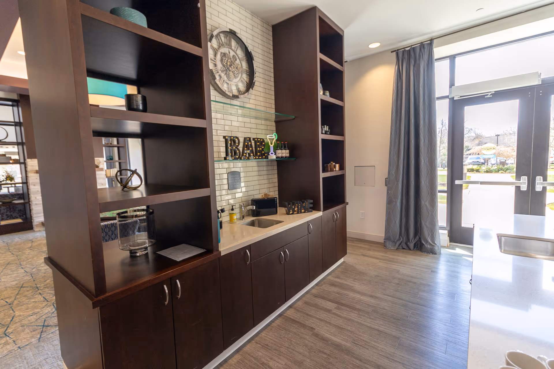 Small bar area with dark wood shelving, a countertop sink, decorative clock and glass doors to the outside.