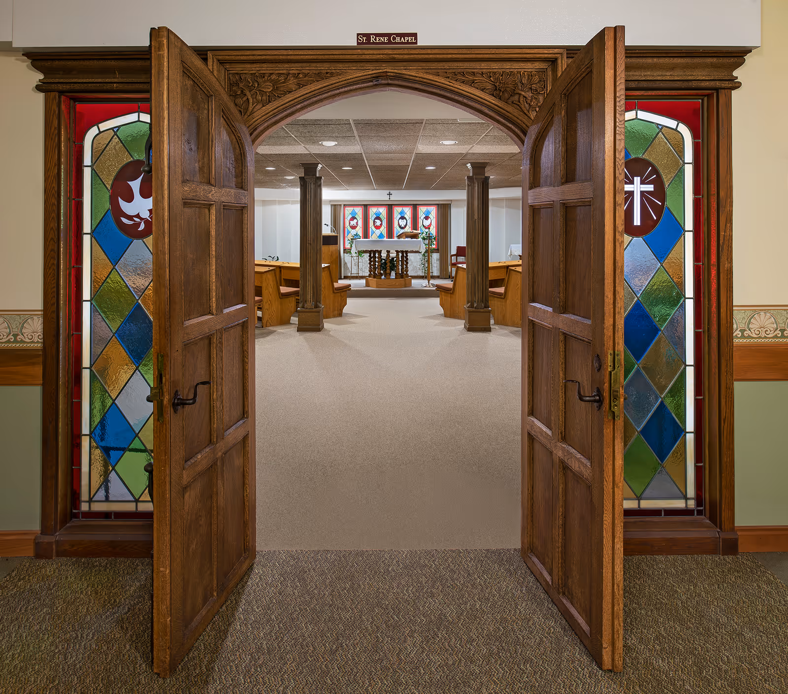 Open wooden double doors with stained glass side panels leading into a small chapel with pews and an altar.