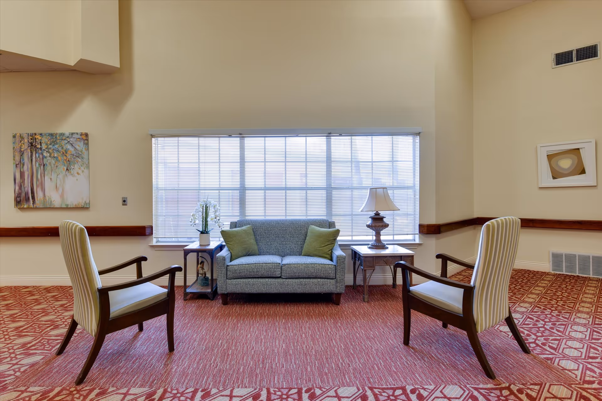 A cozy sitting area in a senior living facility featuring a small gray loveseat with two green pillows, flanked by two wooden side tables. One side table holds a white orchid plant, and the other has a traditional table lamp. Two striped armchairs face the loveseat, all set on a red patterned carpet. The background includes a large window with blinds and light beige walls adorned with two framed artworks.