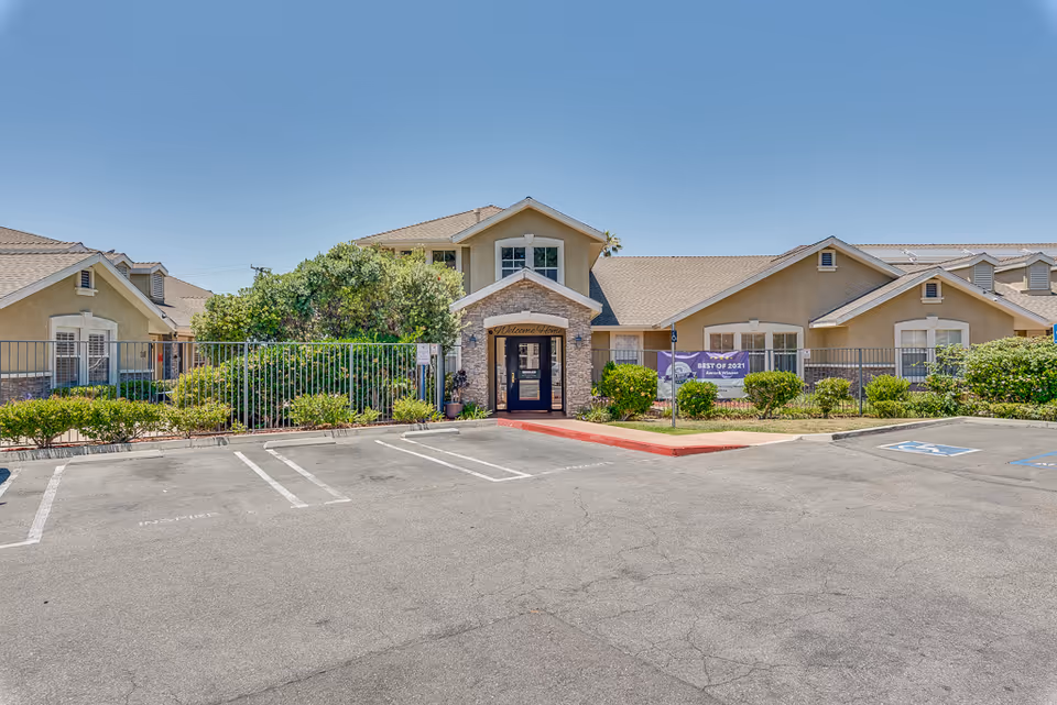 Front entrance of Newport Mesa Senior Living building with parking lot, fence, and landscaping under a clear sky.