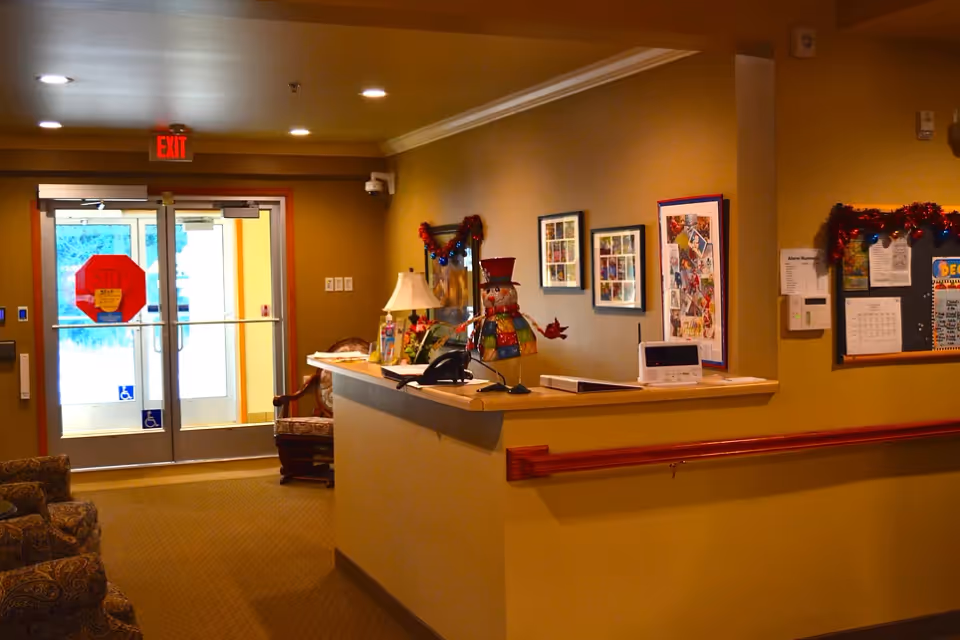 Interior view of a senior living facility reception area with a front desk decorated with a colorful snowman figure, a lamp, and framed pictures on the wall. There are comfortable chairs, a bulletin board with notices, and double glass doors with an exit sign above them.