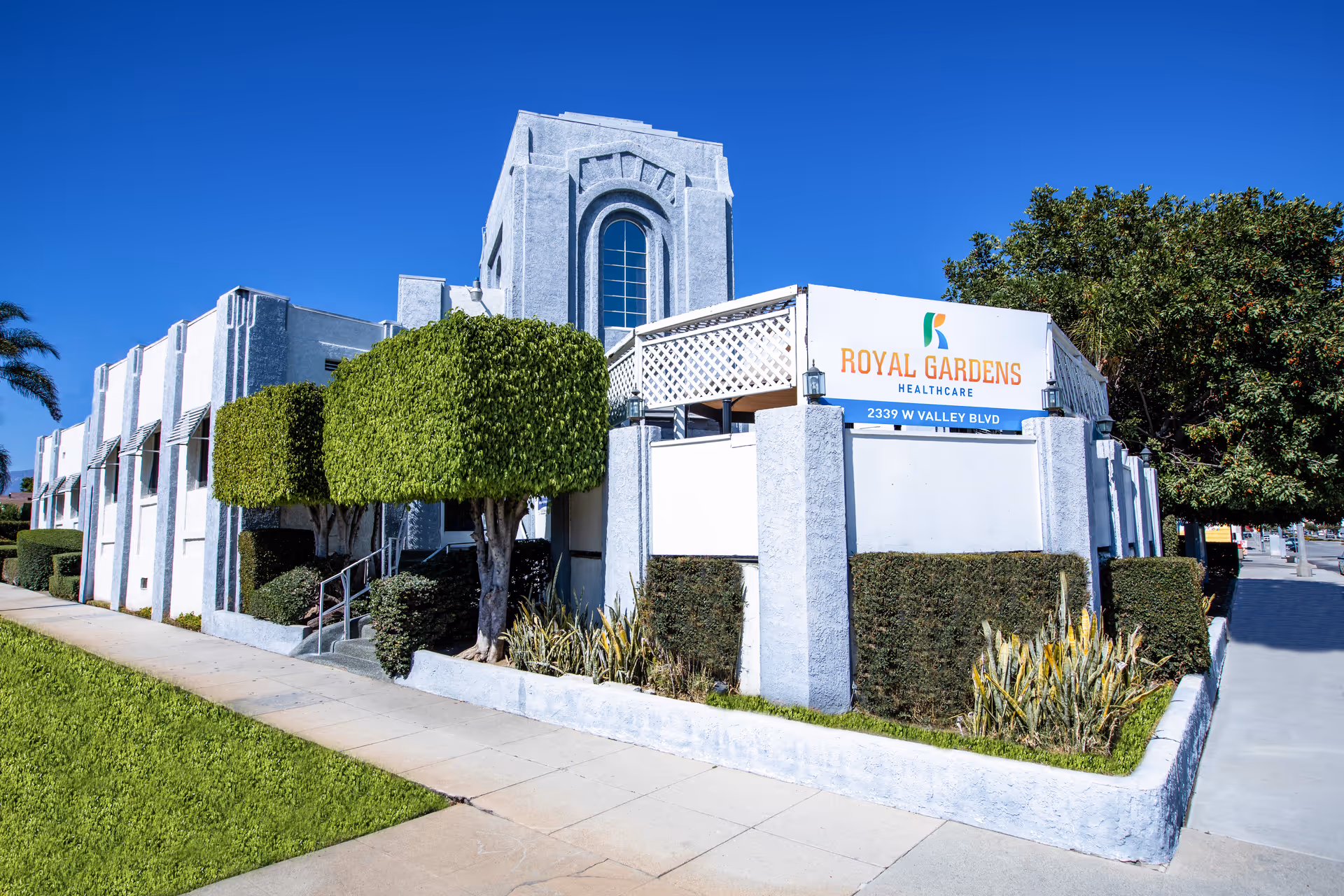 Front exterior of a white Royal Gardens Healthcare building with neatly trimmed trees and a sign reading 'Royal Gardens Healthcare'.