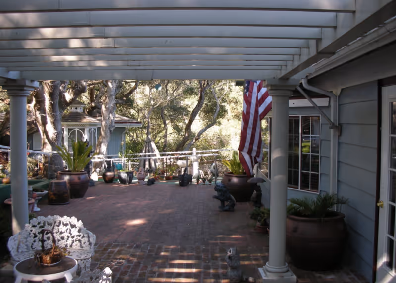 A covered outdoor patio area with brick flooring, white columns, and a white pergola overhead. The patio is decorated with large potted plants, garden statues, and an American flag hanging from one of the columns. Trees and greenery are visible in the background, along with a small structure resembling a gazebo.