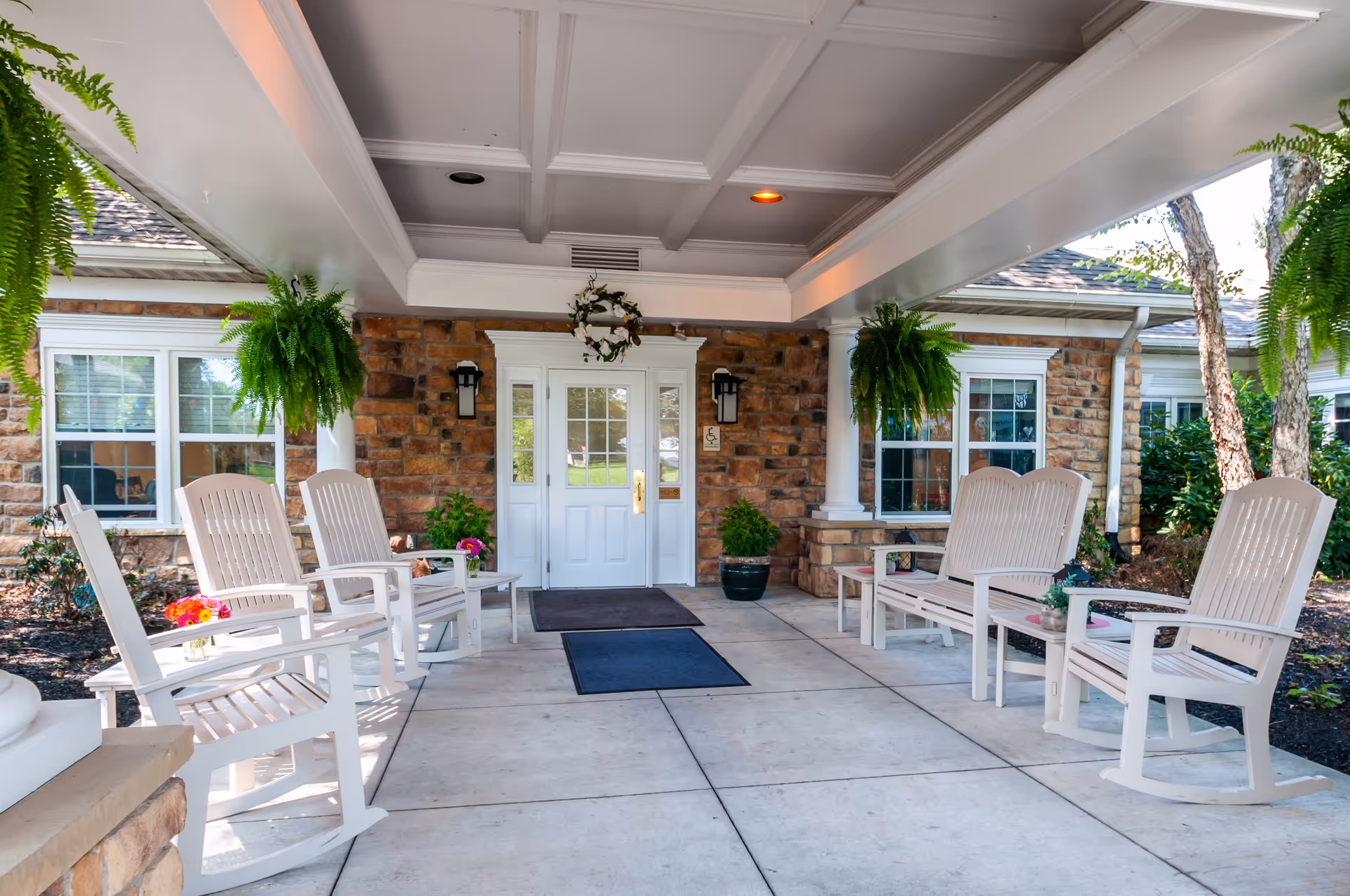 Covered entrance area of a senior living facility with white rocking chairs and benches arranged on both sides. The building has a stone facade with white-framed windows and a white door decorated with a wreath. Green hanging plants and potted plants add greenery to the space.