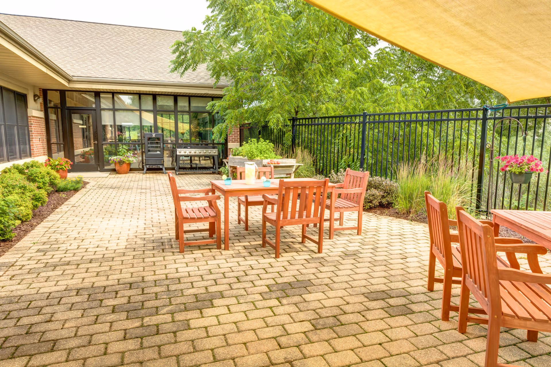 Outdoor patio area with wooden tables and chairs on a paved surface, surrounded by greenery and a black metal fence. There are potted plants and a grill near the building entrance, with a large tree providing shade.