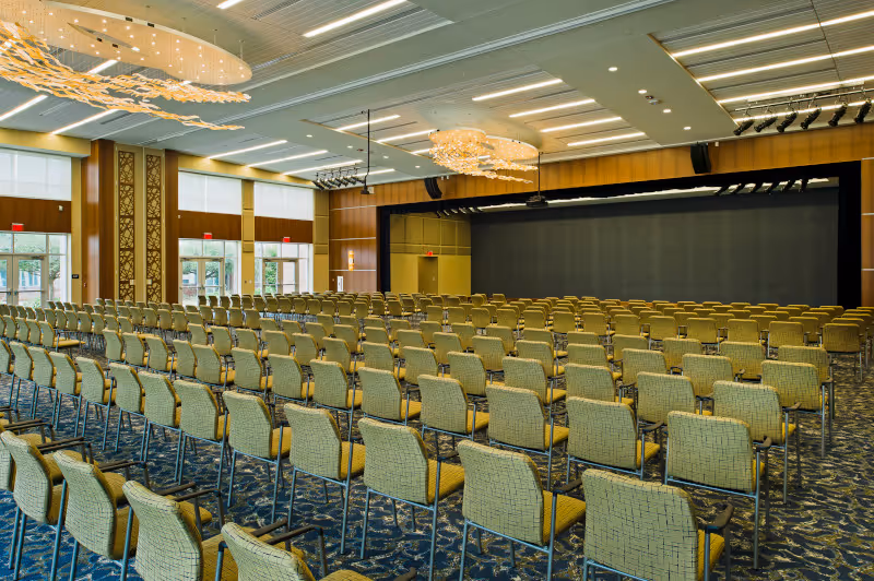 Spacious auditorium with many rows of yellow upholstered chairs facing a stage under decorative ceiling lights.