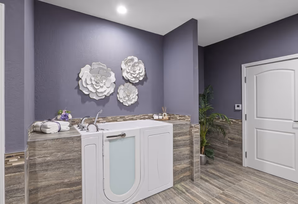 A modern bathroom with a walk-in bathtub featuring a frosted glass door. The walls are painted purple with decorative white flower wall art above the tub. The floor and lower walls are tiled in a gray stone pattern. There is a white door on the right side and a potted plant next to it. Towels and bath accessories are placed on the tub ledge.