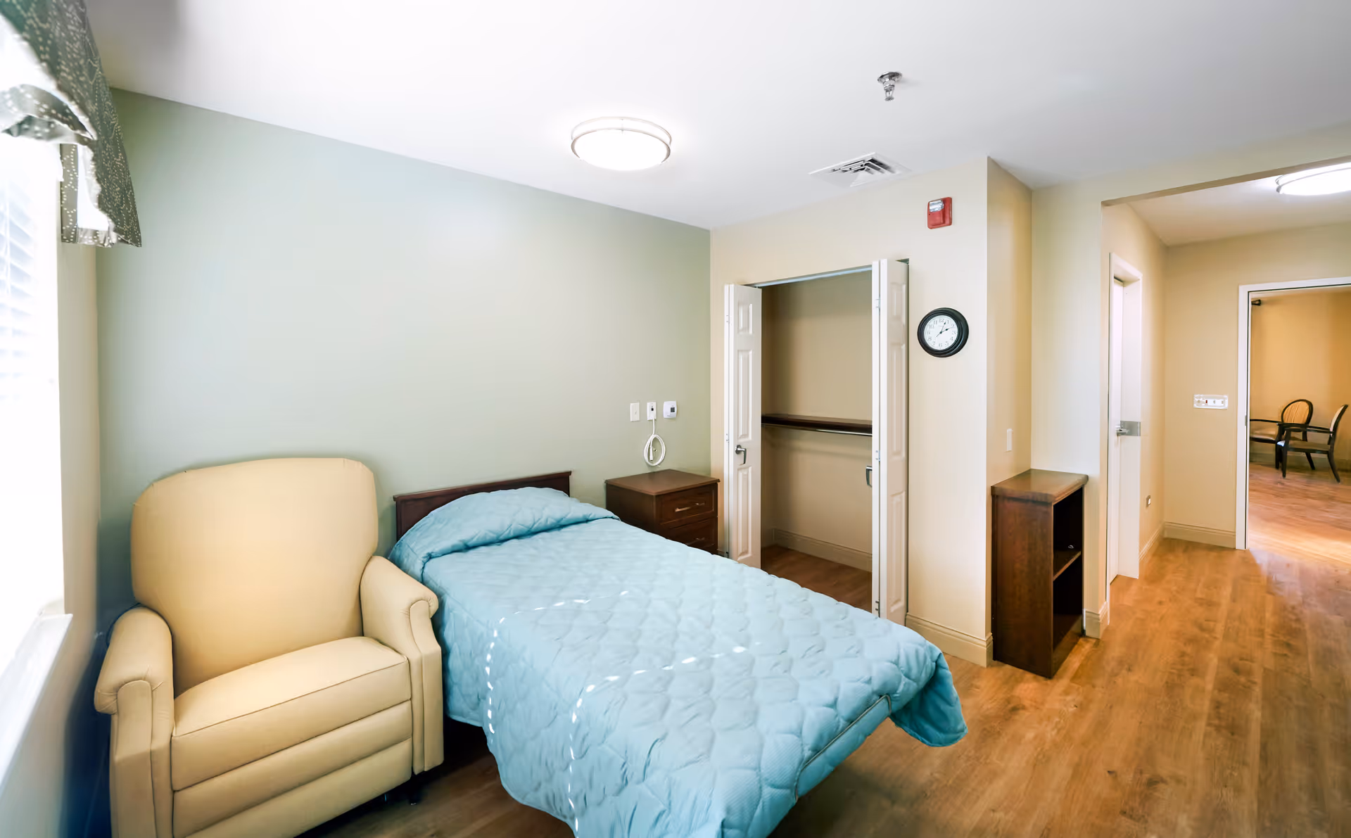 Bright single-occupancy bedroom with a bed, upholstered armchair, open closet and wood flooring.