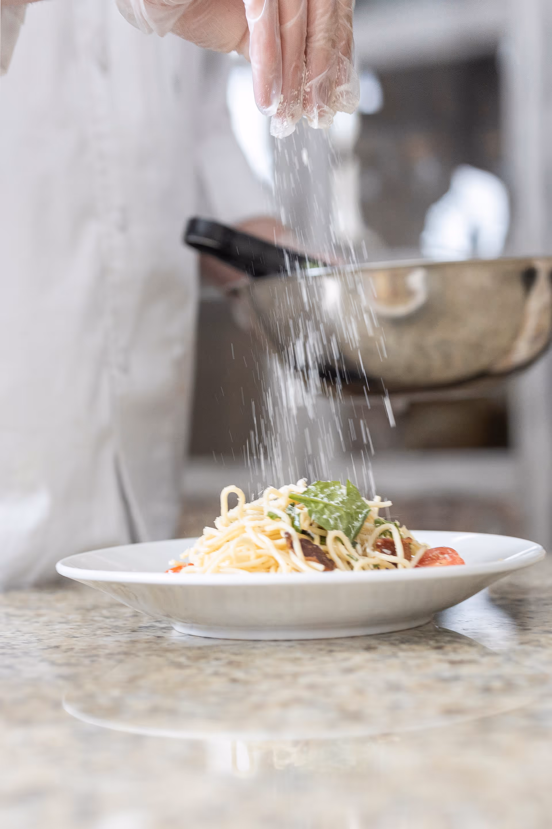 A person wearing a white chef's coat and gloves is sprinkling grated cheese or seasoning over a plate of spaghetti pasta with vegetables on a kitchen counter.