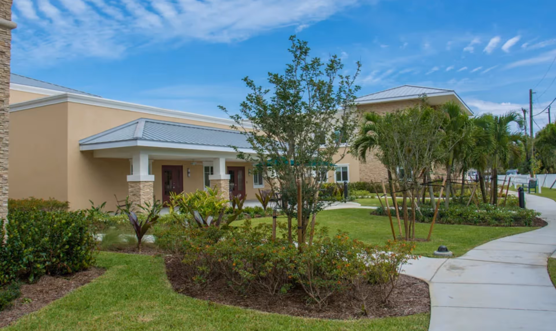 Exterior view of a senior living facility building with beige walls and a metal roof, surrounded by landscaped gardens with various plants, trees, and a curved concrete walkway under a partly cloudy blue sky.