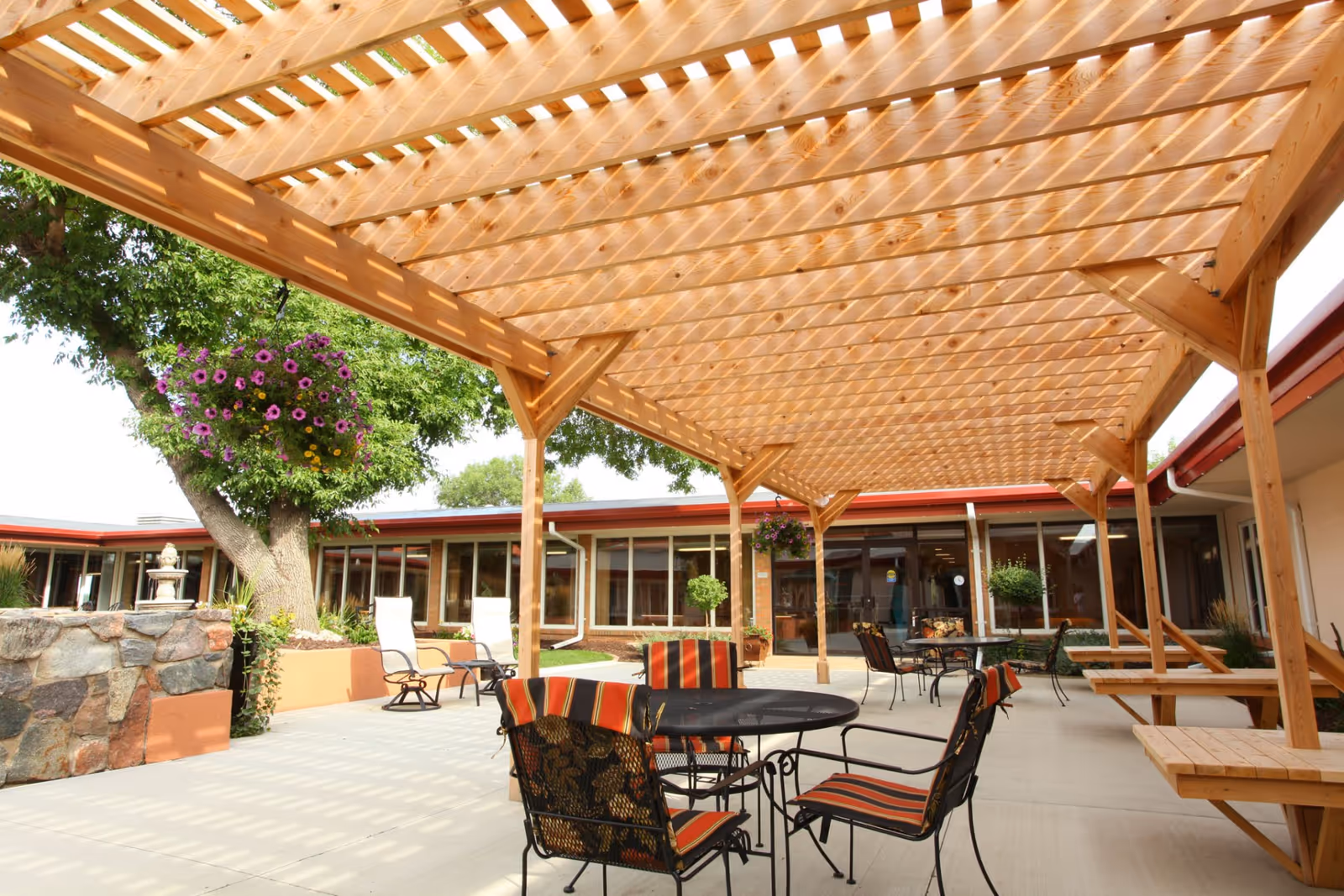 Outdoor patio area with a wooden pergola overhead casting shadows on the concrete floor. There are several metal tables and chairs with striped cushions, hanging flower baskets, and a stone water fountain near a tree. The patio is surrounded by a building with large windows.