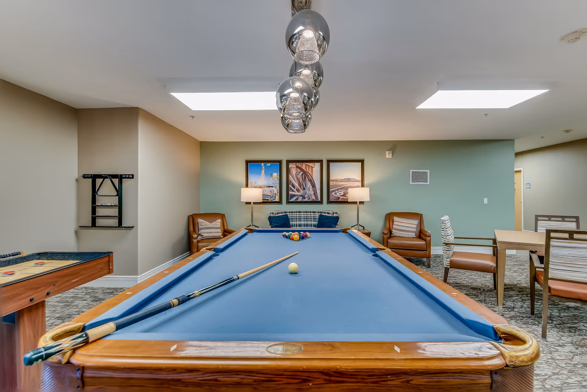 A recreational room with a blue felt pool table in the center, two pool cues resting on it, and a set of billiard balls arranged at the far end. The room has beige walls with a green accent wall, two brown leather armchairs, a plaid sofa, two floor lamps, framed pictures on the wall, a shuffleboard table to the left, and a dining table with chairs to the right. Overhead, there are modern pendant lights and skylights providing natural light.