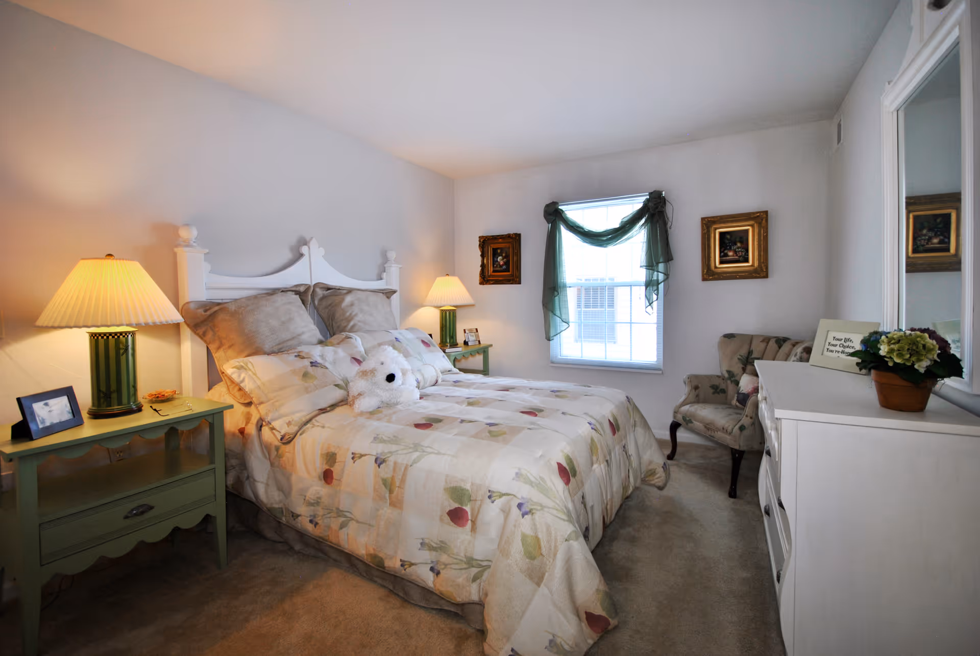 A cozy bedroom with a white wooden bed frame, floral patterned bedding, and a stuffed white teddy bear on the bed. There are two green nightstands with lamps on either side of the bed. A window with green sheer curtains is centered on the far wall, flanked by two framed paintings. To the right, there is a floral upholstered armchair and a white dresser with a potted plant and a small framed sign on top. The room has beige carpeting and light-colored walls.