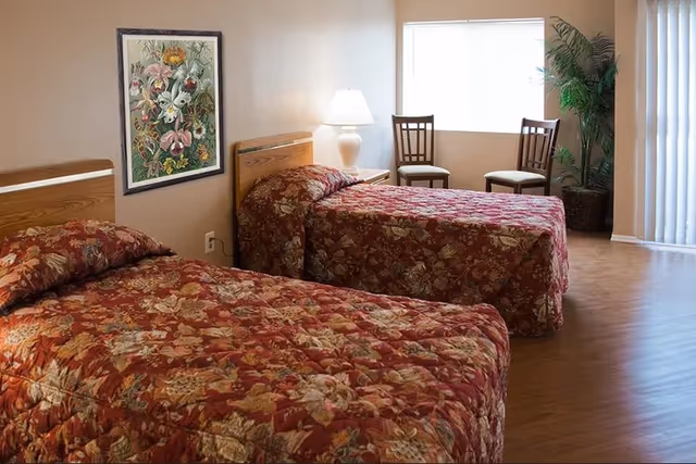 A bedroom with two single beds covered in red floral bedspreads. Between the beds is a nightstand with a white lamp. On the wall above one bed is a framed floral painting. Near a window with blinds are two wooden chairs and a large potted plant. The floor is wooden, and there is a sliding glass door with vertical blinds.