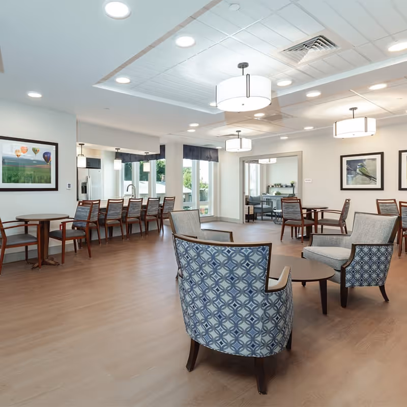 A bright and spacious senior living community common area with wooden flooring, several seating arrangements including patterned armchairs around a small round table, and multiple wooden tables with chairs. The room features large windows allowing natural light, modern ceiling lights, framed artwork on the walls, and a kitchen area with bar stools in the background.