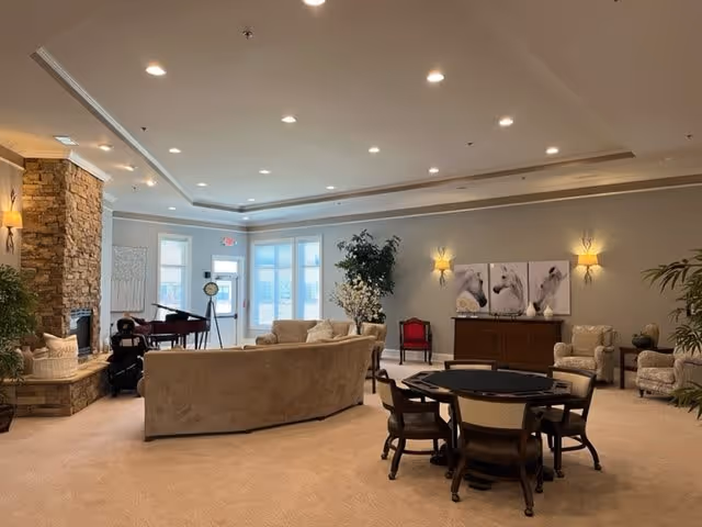 A spacious and well-lit living room area in an assisted living facility featuring beige carpeting, a curved beige sofa, several armchairs, a round table with chairs, a stone fireplace, a piano, and wall art depicting horses. The room has recessed lighting and large windows allowing natural light.