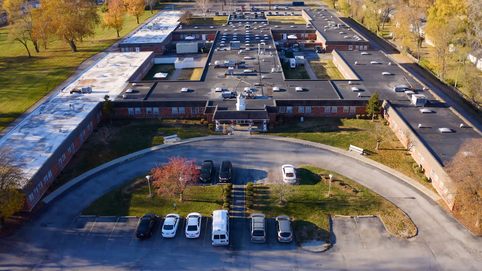 Aerial view of St Sophia Health & Rehabilitation Center showing a large, single-story building with a flat roof and multiple wings arranged in a U-shape. The building is surrounded by a circular driveway with parked cars and green lawns with trees displaying autumn foliage.