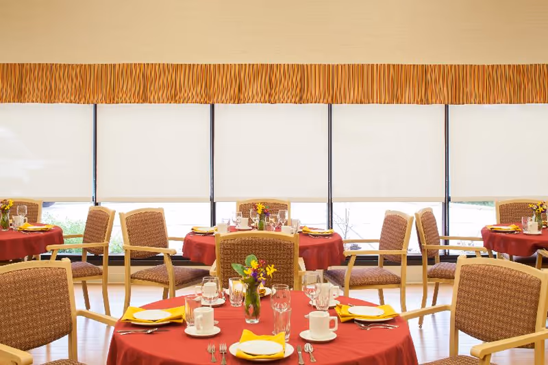 Dining room with round tables covered in red tablecloths, place settings and flower vases, surrounded by wooden chairs in front of large windows with blinds.
