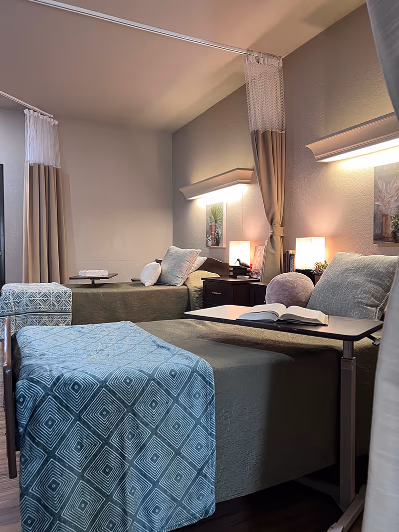 A cozy room with two single beds, each with green bedspreads and decorative pillows. One bed has a blue patterned throw blanket and an adjustable overbed table with an open book. The room has soft lighting from wall-mounted lamps and table lamps on a nightstand between the beds. Beige curtains hang from ceiling-mounted tracks, and framed artwork decorates the walls.
