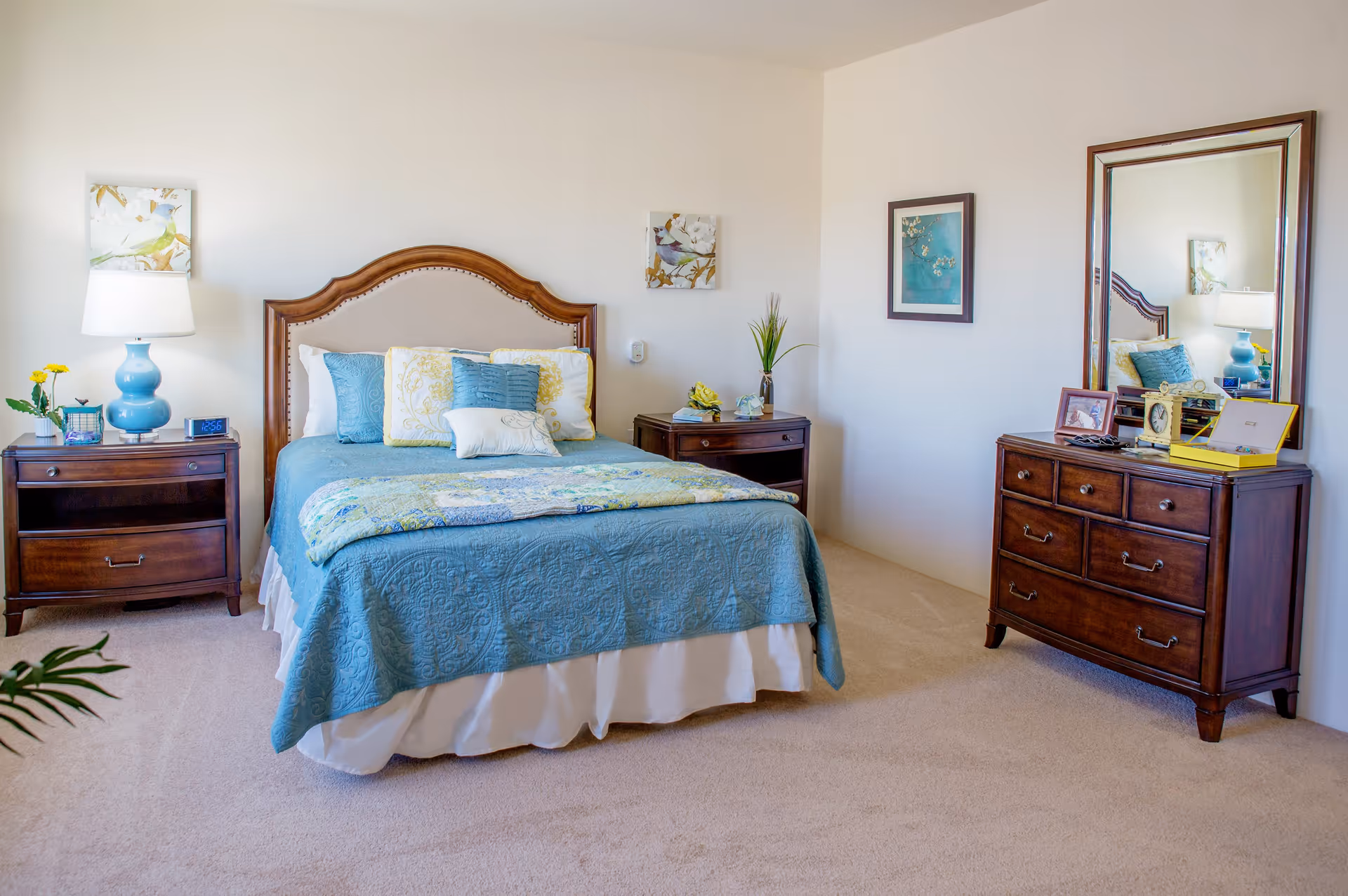 A neatly made bed with a blue quilt and multiple pillows in a bedroom. The room features two wooden nightstands with a lamp, clock, and decorative items. There is a wooden dresser with a large mirror, framed pictures, and other small decor. The walls are light-colored with framed artwork, and the floor is carpeted.