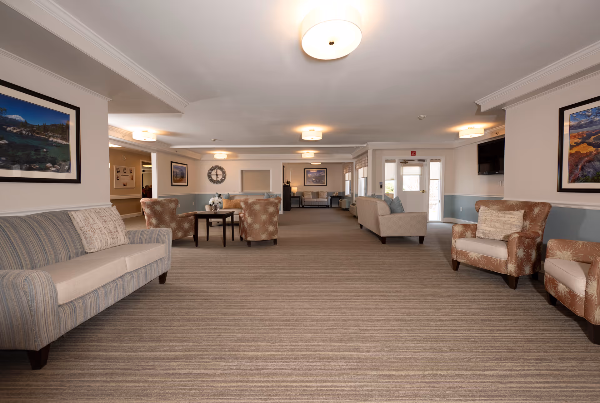 A spacious senior living facility common area with multiple seating arrangements including sofas and armchairs. The room has neutral-toned carpeting, soft lighting from ceiling fixtures, framed landscape artwork on the walls, and a wall-mounted television. There are windows and a door at the far end of the room allowing natural light to enter.