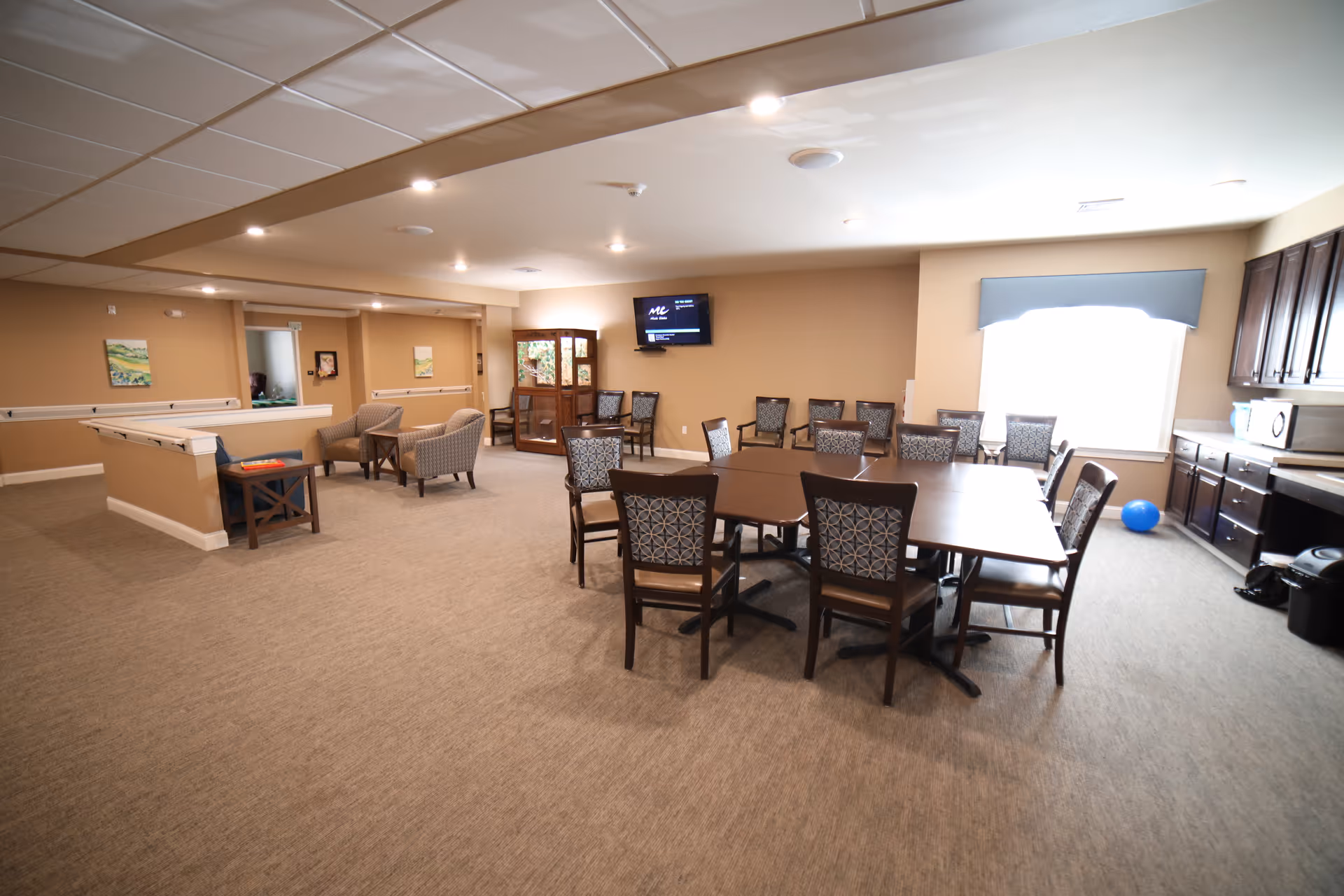 A spacious common area in a senior living facility with a large table surrounded by chairs in the center, additional chairs lined up against the wall, a television mounted on the wall, a cabinet with glass doors, and a seating area with armchairs. The room has beige walls, carpeted floor, and a window with a blue valance letting in natural light.
