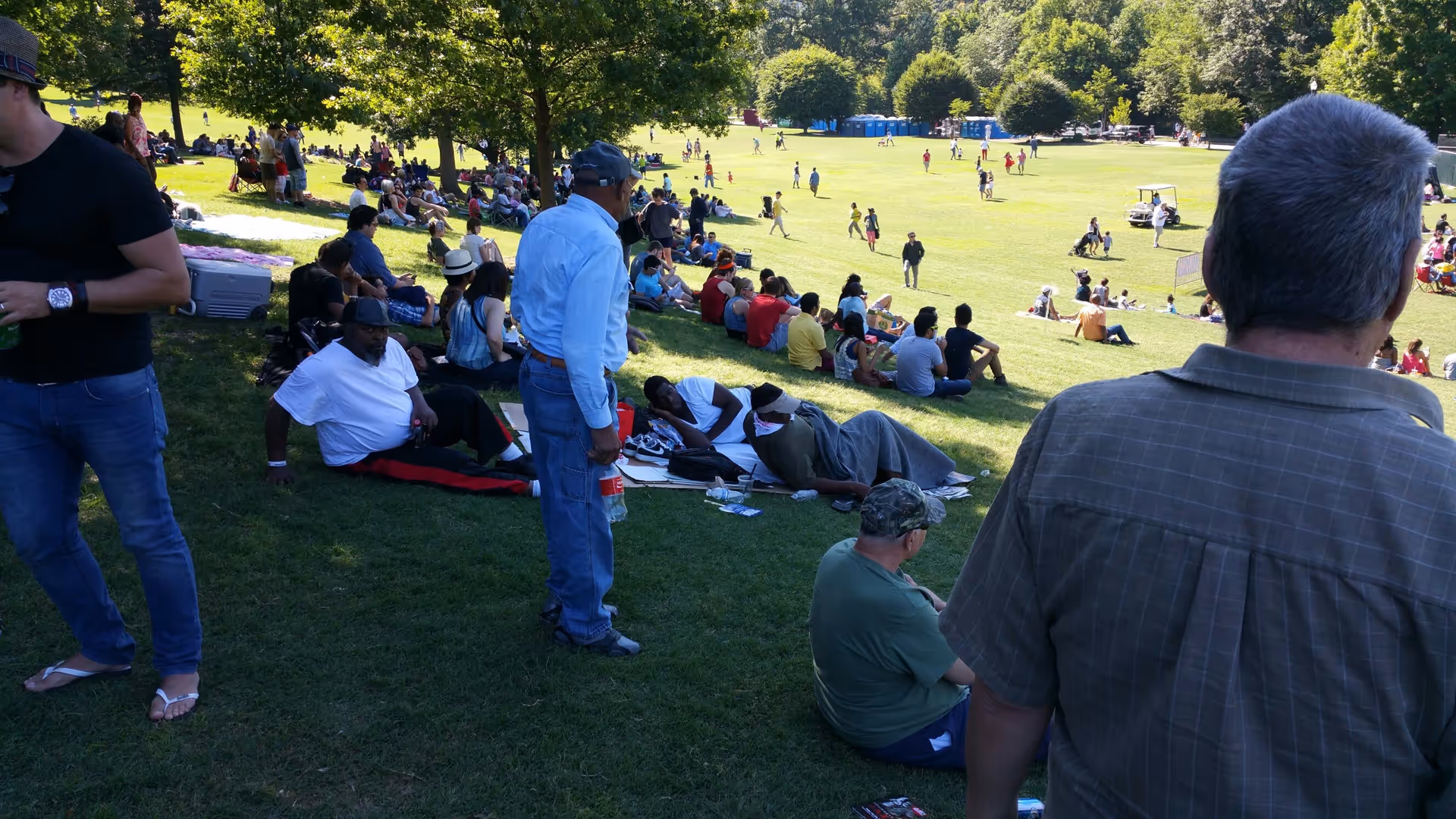 A large group of people sitting and relaxing on a grassy hill in a park on a sunny day. Some people are sitting on blankets or chairs, while others are standing or walking. Trees provide shade in the background, and there are portable toilets visible in the distance.