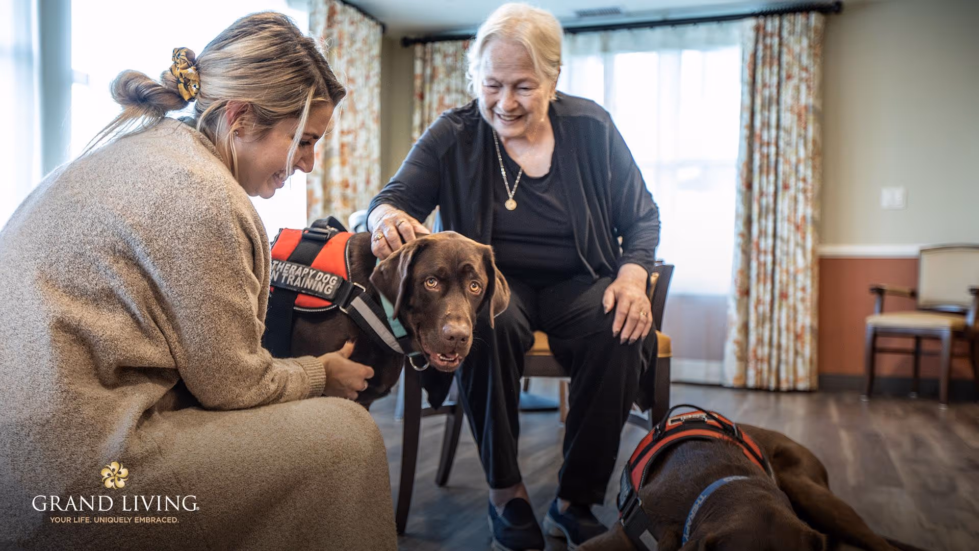 Two women in a communal living room smile and pet therapy dogs, one wearing a 'Therapy Dog in Training' vest.
