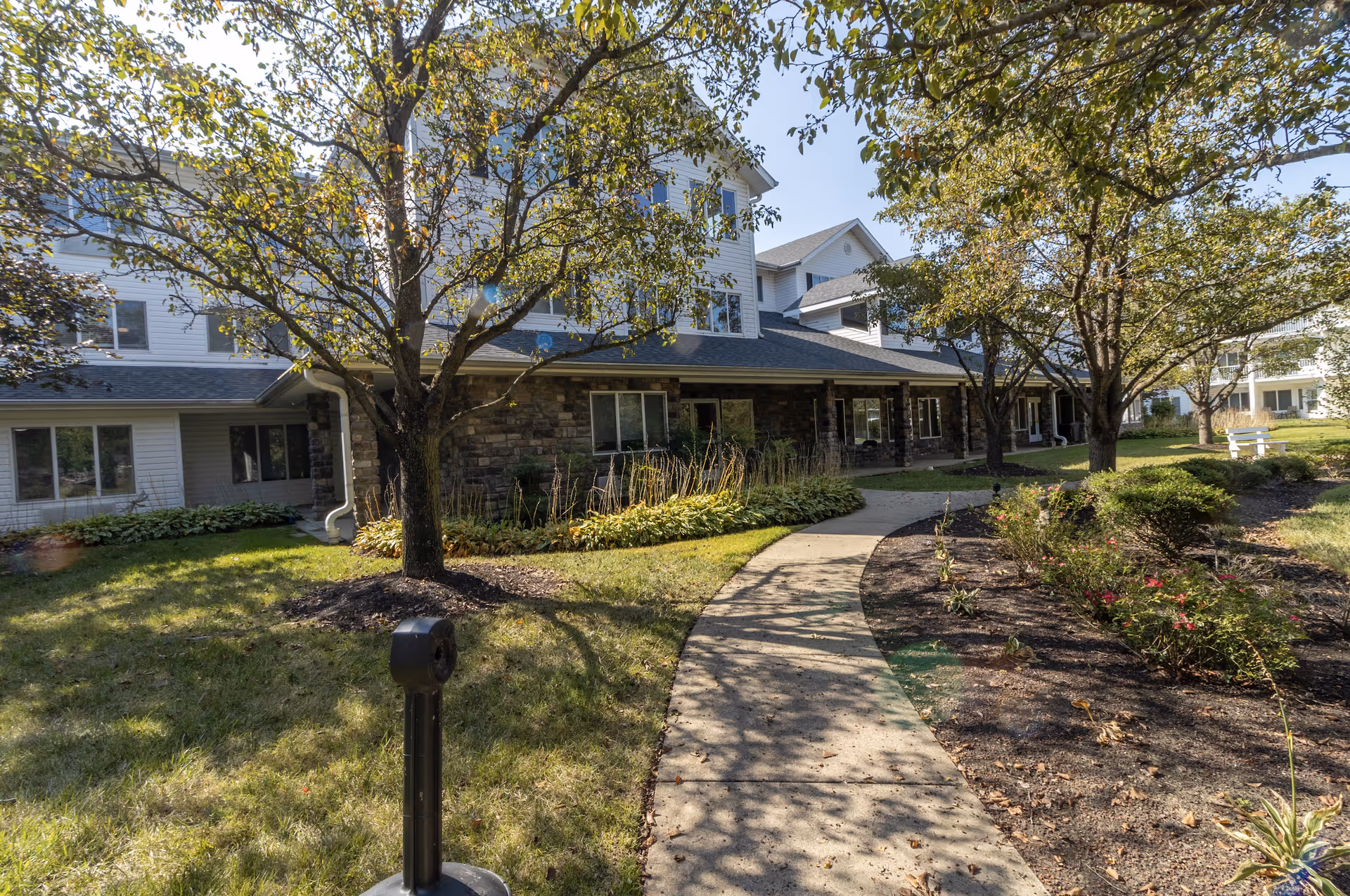 Outdoor view of a senior living facility with a paved walkway leading through a landscaped garden area with trees and shrubs. The building has multiple stories with white siding and stone accents on the lower level. The scene is sunny with shadows cast by the trees.