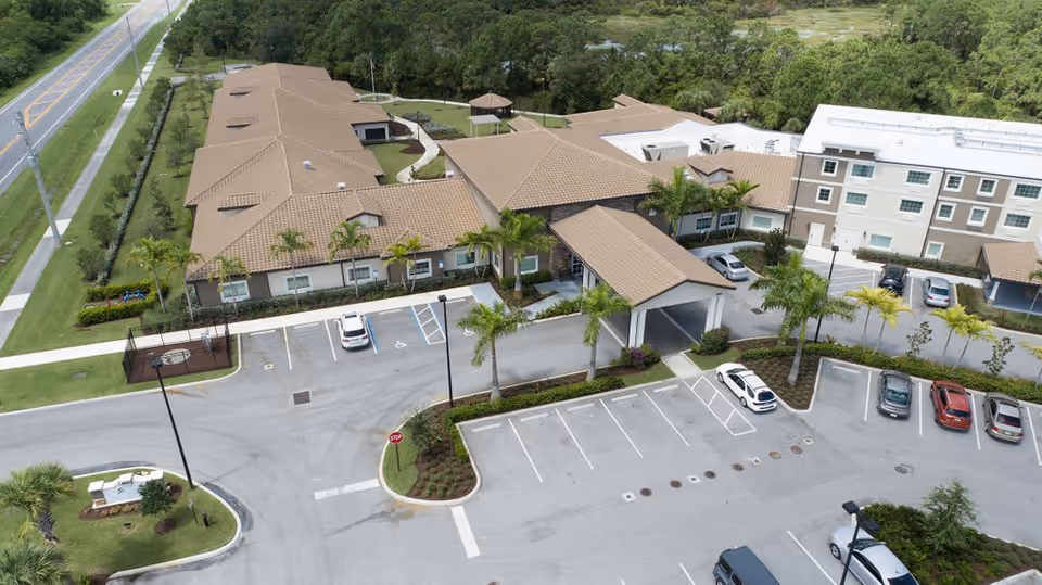 Aerial view of Discovery Village At Stuart senior living facility showing multiple connected buildings with tan roofs, a covered entrance, palm trees, parking lots with several cars, and surrounding greenery along a nearby road.