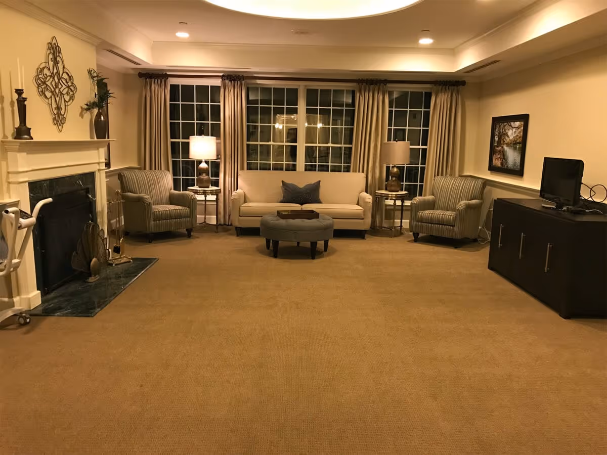 A cozy living room with beige carpet, a beige sofa with a dark cushion, two striped armchairs, two side tables with lamps, a dark ottoman in the center, a fireplace on the left, and a TV on a dark cabinet on the right. Large windows with beige curtains are behind the sofa.