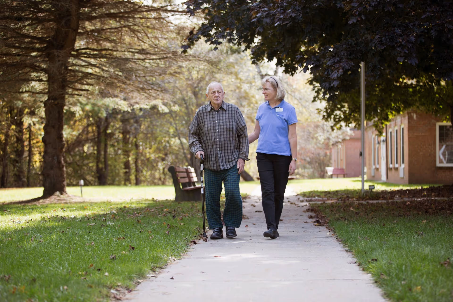 An elderly man walking with a cane is accompanied by a female caregiver wearing a blue shirt and black pants. They are walking on a paved path outdoors, surrounded by green grass, trees, and a building in the background.