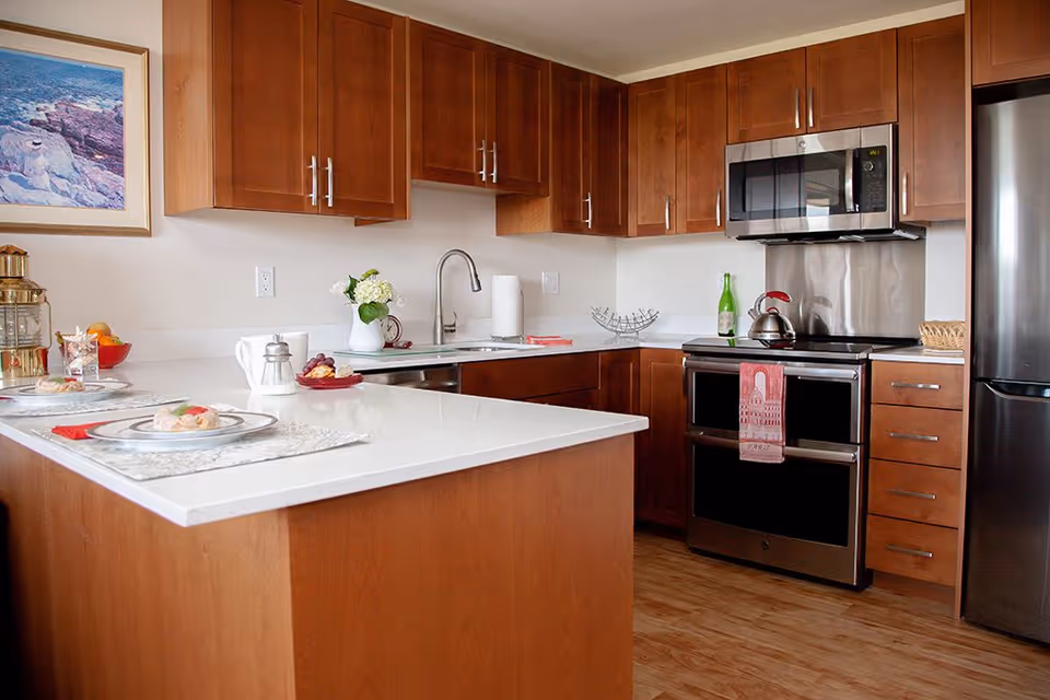 A modern kitchen with wooden cabinets, a white countertop island with place settings, a stainless steel refrigerator, oven, and microwave. There is a sink with a faucet, a kettle on the stove, a green bottle, and a framed picture on the wall.