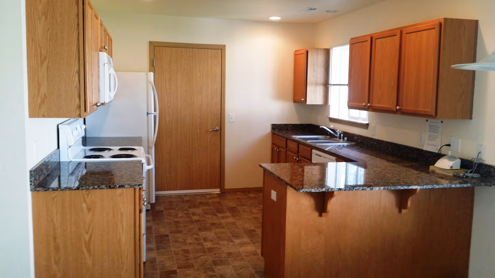 A kitchen with wooden cabinets, granite countertops, a white electric stove, microwave, refrigerator, and a double sink under a window. The floor has a brown tile pattern, and there is a wooden door at the end of the kitchen.