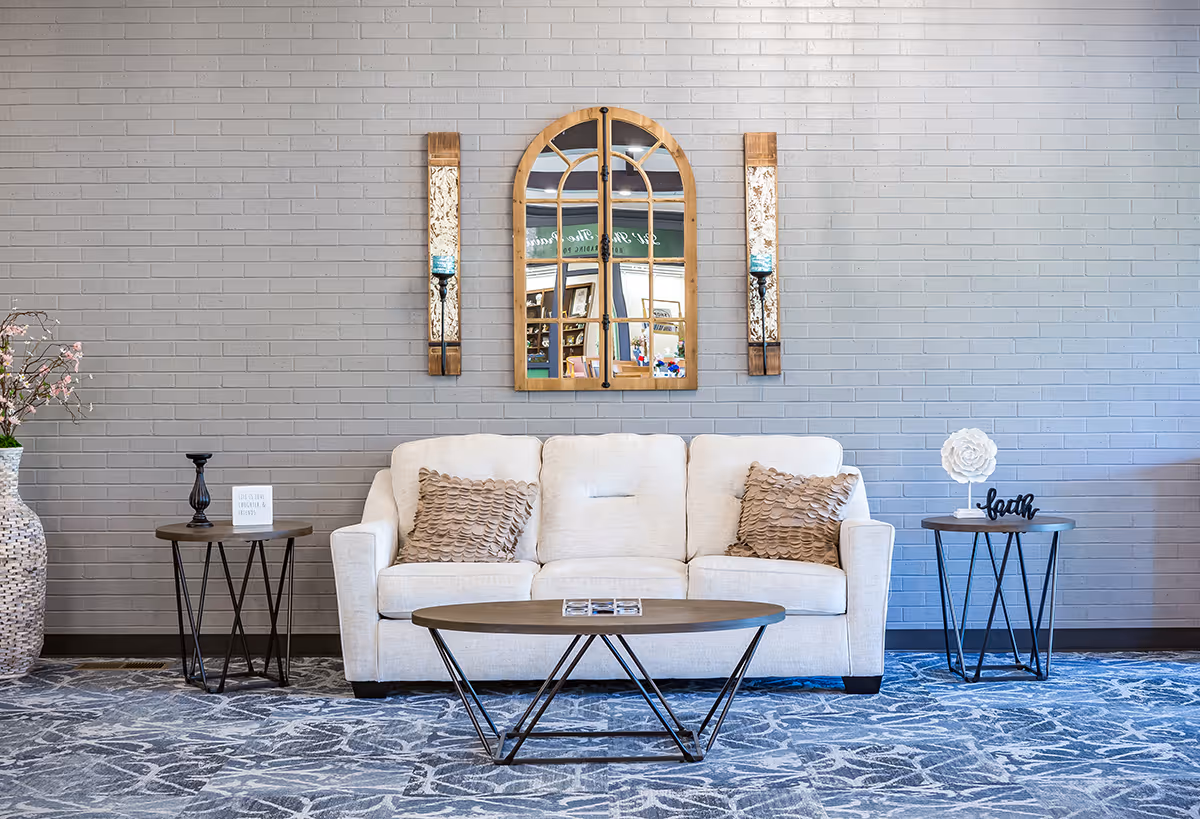 A cozy living room area with a light beige sofa adorned with two textured pillows. In front of the sofa is a modern oval coffee table with a geometric metal base. On either side of the sofa are round side tables with decorative items, including a black candlestick, a small sign, and a white flower sculpture with the word 'faith'. The background features a gray brick wall with a large arched mirror and two vertical wall sconces with candles. A large vase with pink flowers is placed on the left side. The floor is covered with a patterned blue and gray carpet.