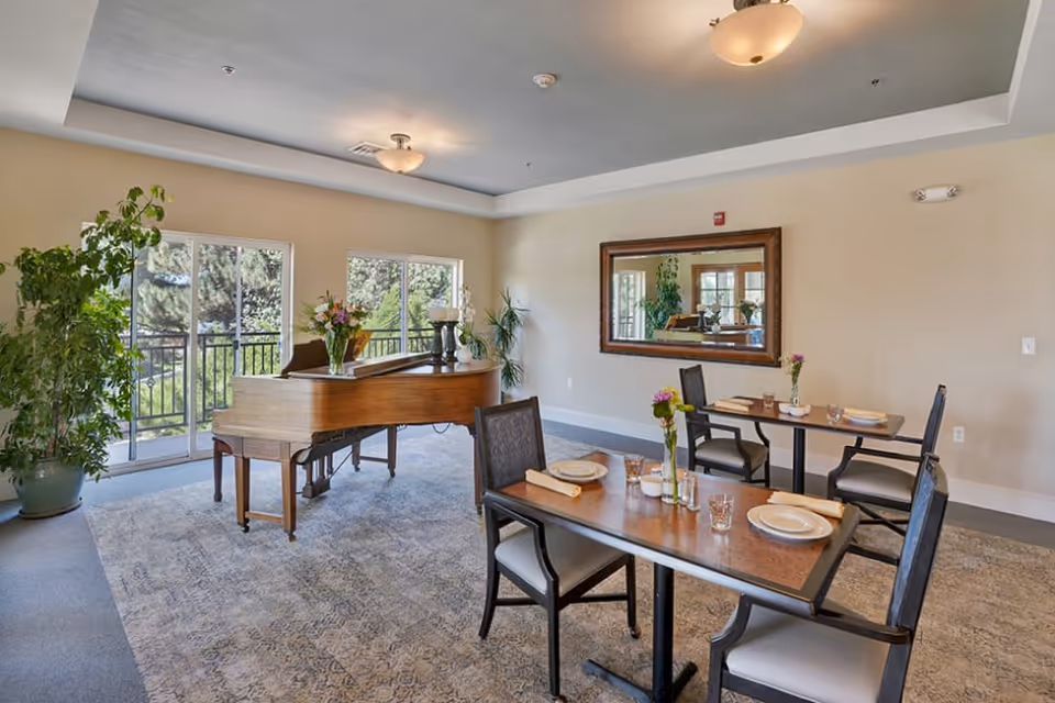 A bright room with two dining tables set with plates, glasses, and napkins. A grand piano with a flower vase on top is positioned near large windows that let in natural light. There are several potted plants and a large mirror on the wall reflecting part of the room.