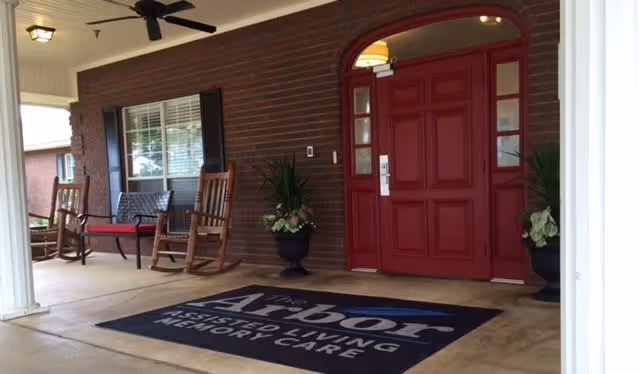 Covered front porch area with a red door, two wooden rocking chairs, a bench, potted plants, ceiling fan, and a floor mat that reads 'Arbor Assisted Living Memory Care'.
