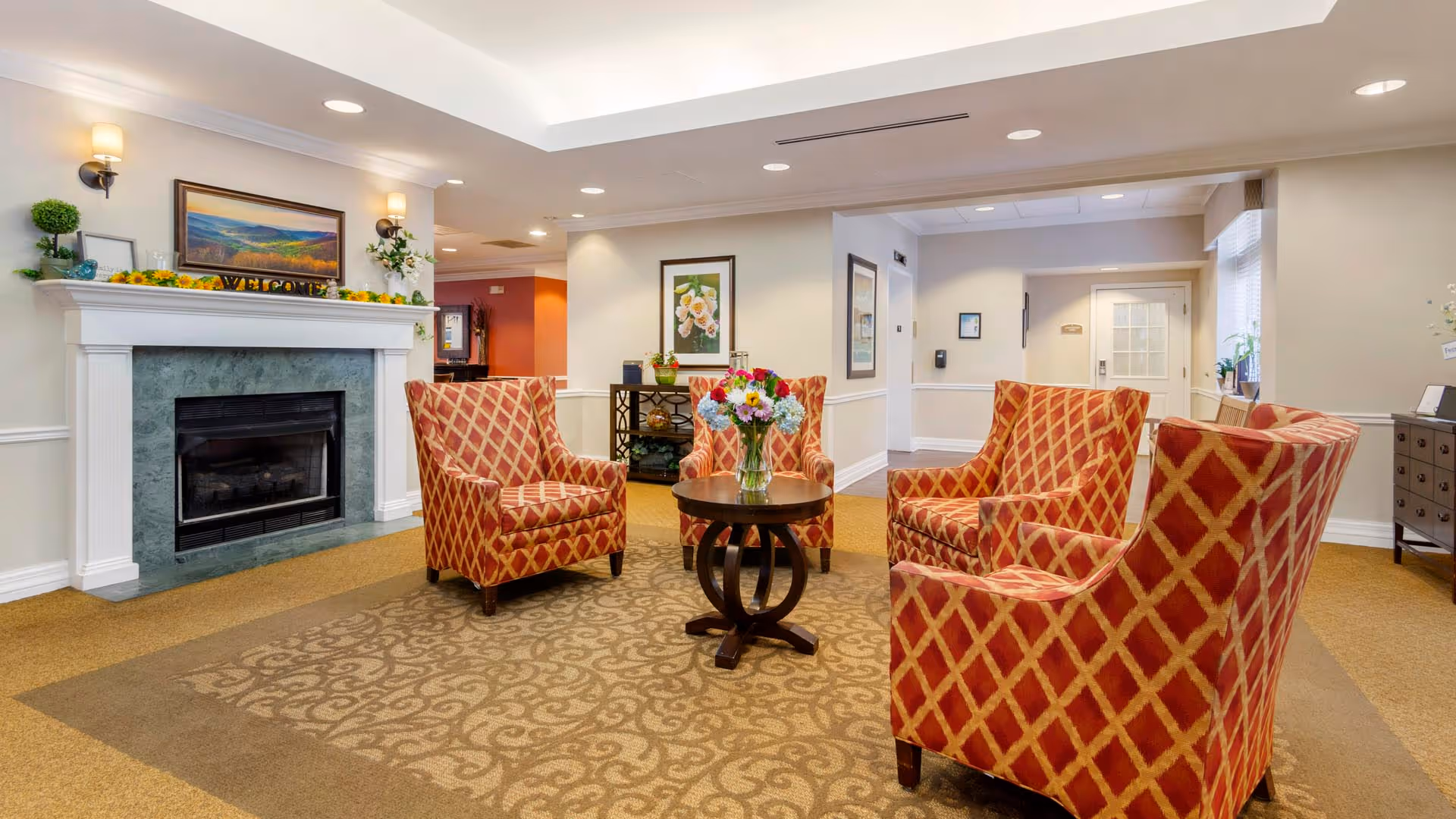 A cozy common area with four red and gold patterned armchairs arranged around a small round wooden table with a vase of colorful flowers. The room features a fireplace with a green marble surround and a white mantel decorated with plants, a framed landscape painting, and a welcome sign. The walls are light-colored with framed artwork, and the space is well-lit with recessed ceiling lights and wall sconces.