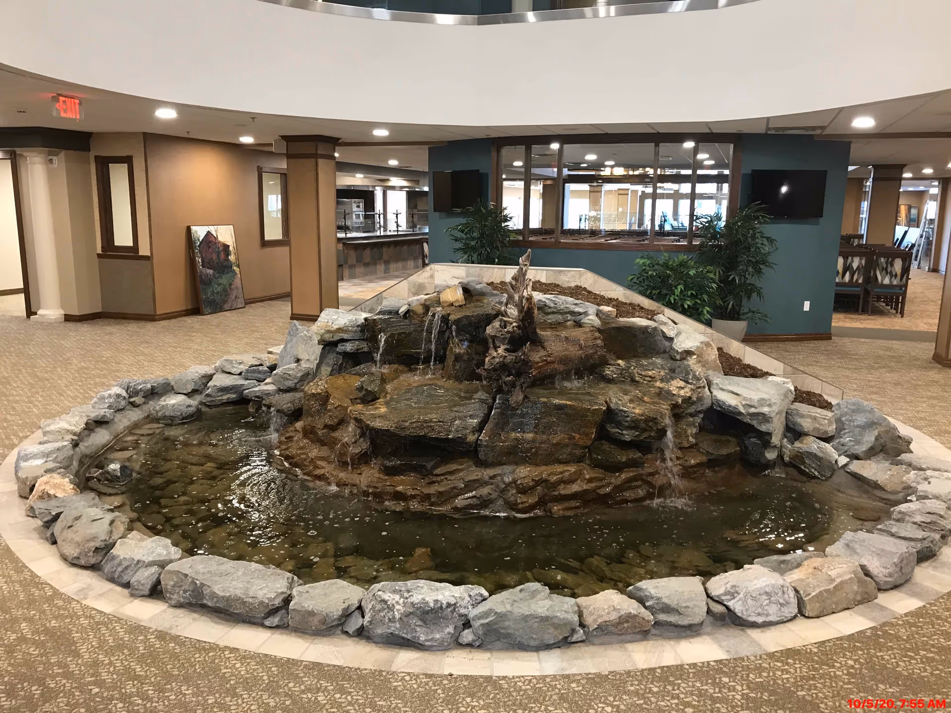 Indoor water feature with rocks and a small waterfall in the center of a carpeted common area with plants, a television mounted on the wall, and seating areas in the background.