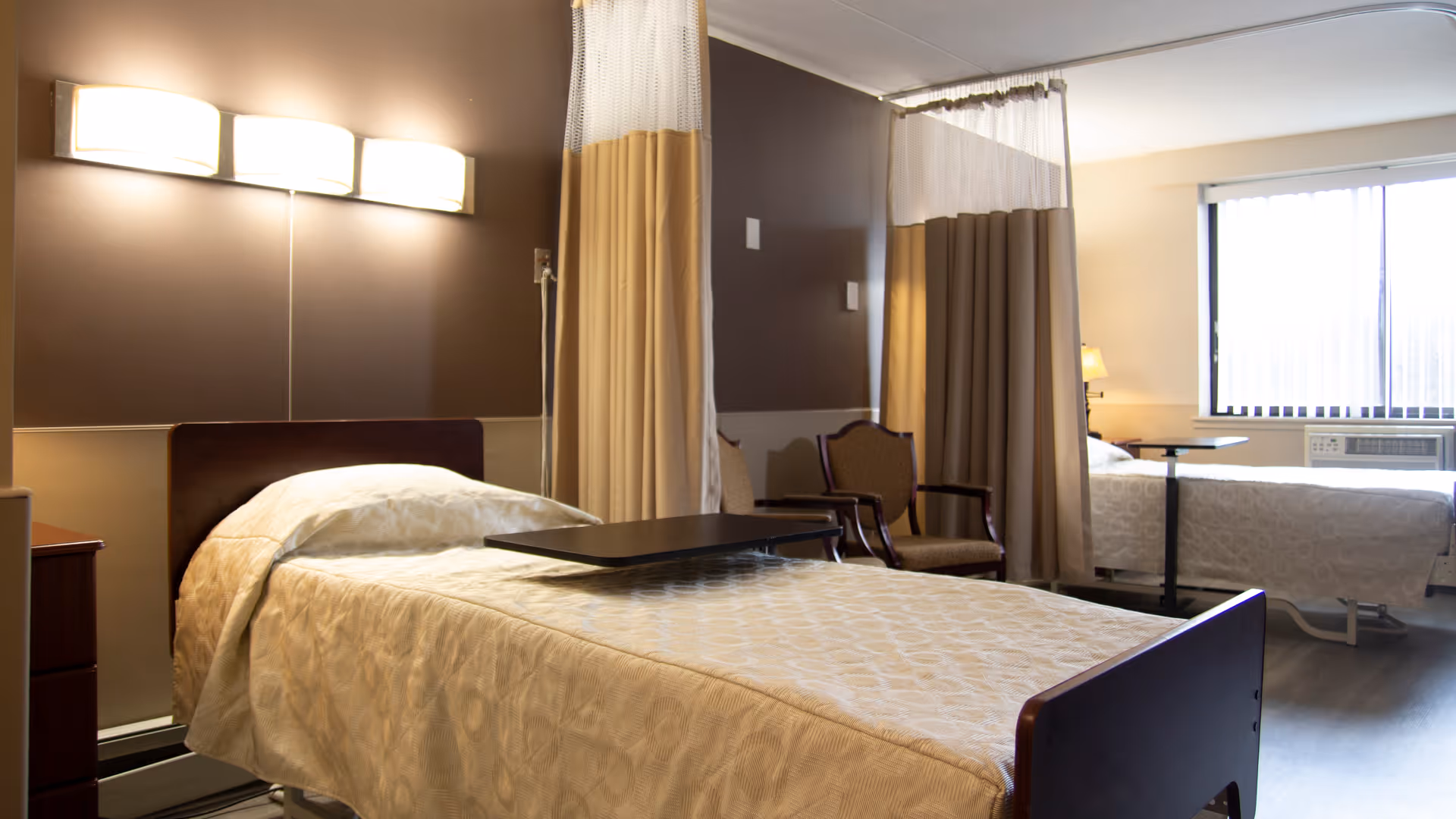 A well-lit room with two hospital-style beds separated by a privacy curtain. Each bed has a beige patterned bedspread and an overbed table. There are two chairs positioned near the curtain, and a window with vertical blinds allows natural light into the room. The walls are painted in neutral tones, and a wall-mounted light fixture is above one of the beds.