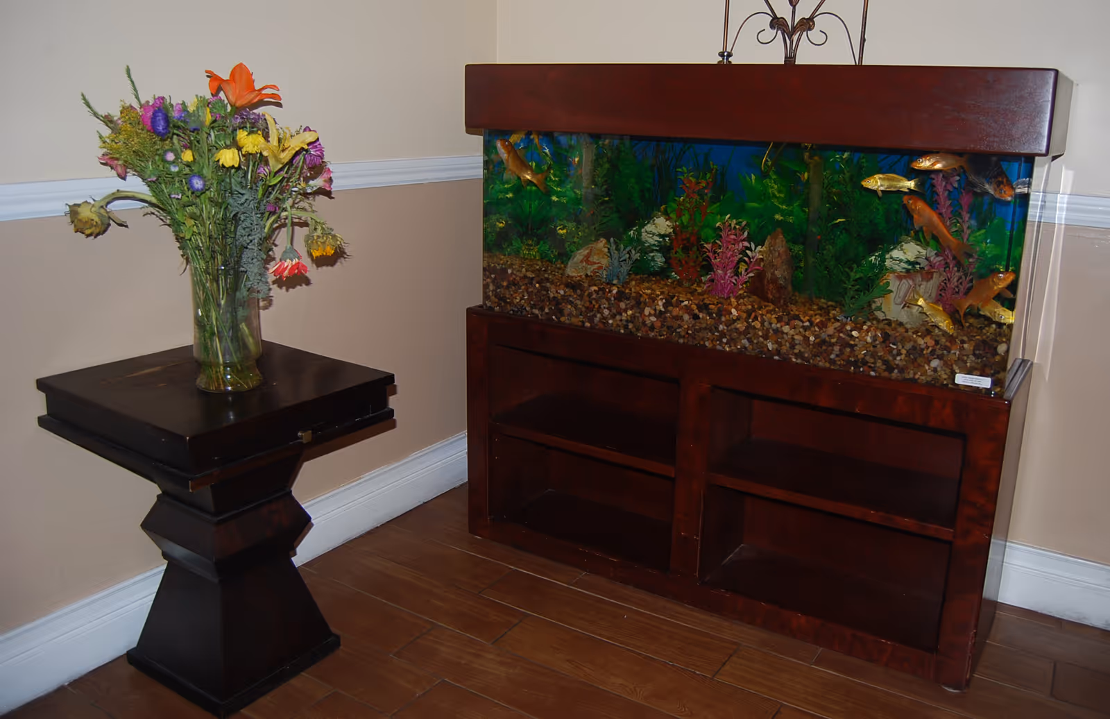 A corner of a room featuring a large fish tank with colorful fish and aquatic plants inside, placed on a dark wooden stand. Next to the fish tank is a small dark wooden table with a vase holding a bouquet of various flowers, including orange, yellow, purple, and pink blooms. The room has light beige walls with white trim and wooden flooring.