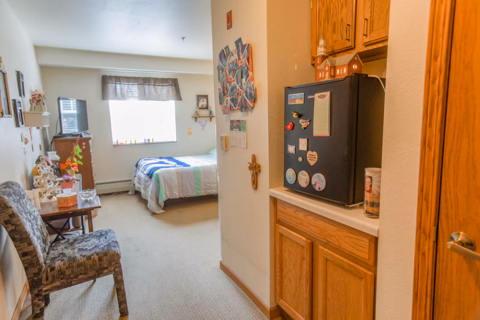 A small assisted-living studio with a bed by a window, a chair and side table, and a kitchenette area with a small refrigerator and cabinets.