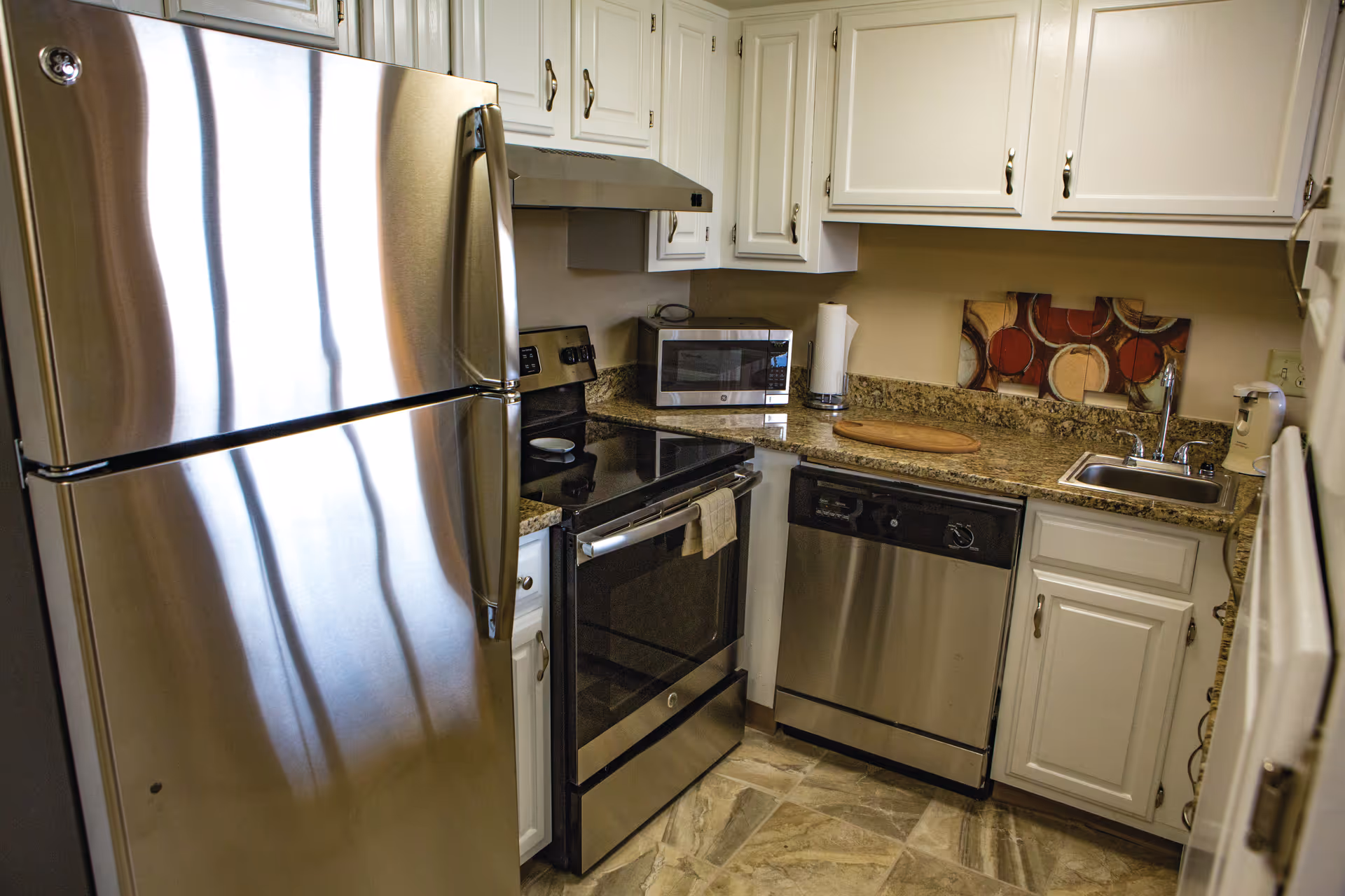 A compact kitchen featuring stainless steel appliances including a refrigerator, oven, microwave, and dishwasher. The kitchen has white cabinets, granite countertops, a small sink, a paper towel holder, a cutting board, and a decorative wall art piece with circular patterns.