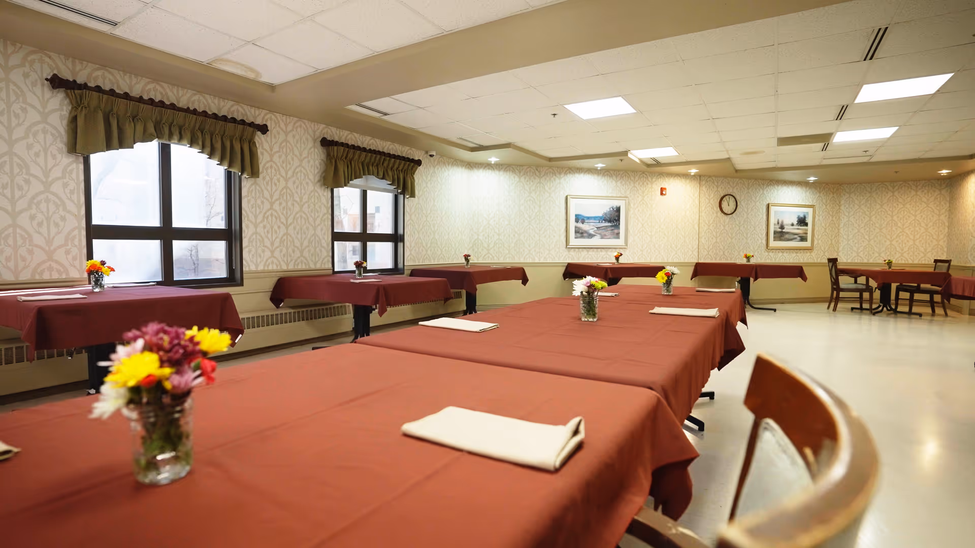 A dining room with multiple tables covered in red tablecloths, each adorned with a small vase of colorful flowers and a folded napkin. The room has patterned wallpaper, several windows with green valances, framed landscape paintings on the walls, and a clock. The floor is light-colored and the ceiling has recessed lighting.