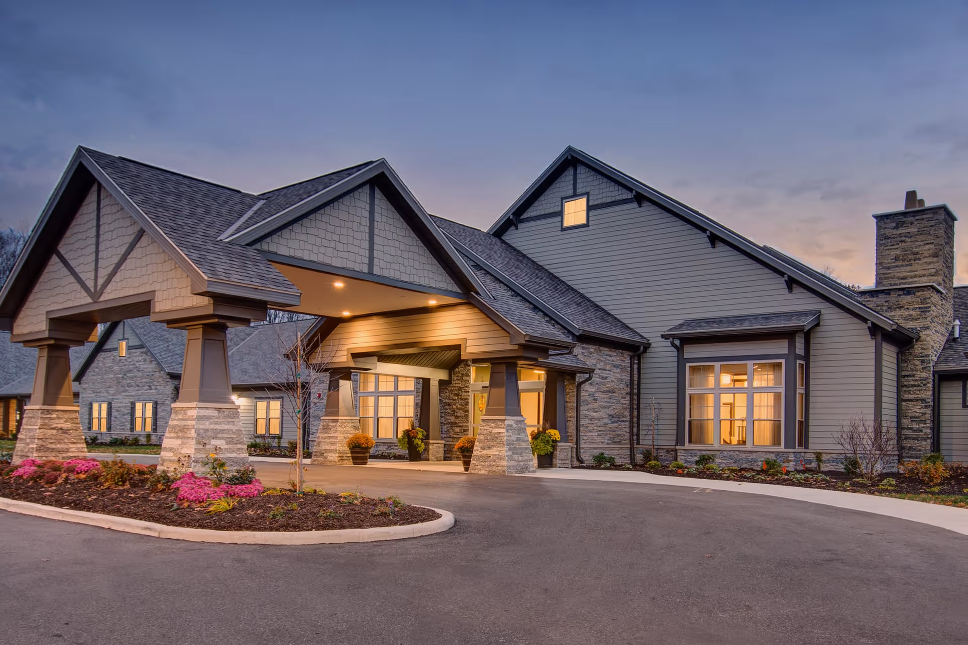 Exterior view of a senior living facility building at dusk with warm lights glowing from the windows, a covered entrance supported by stone pillars, and landscaped flower beds in front.
