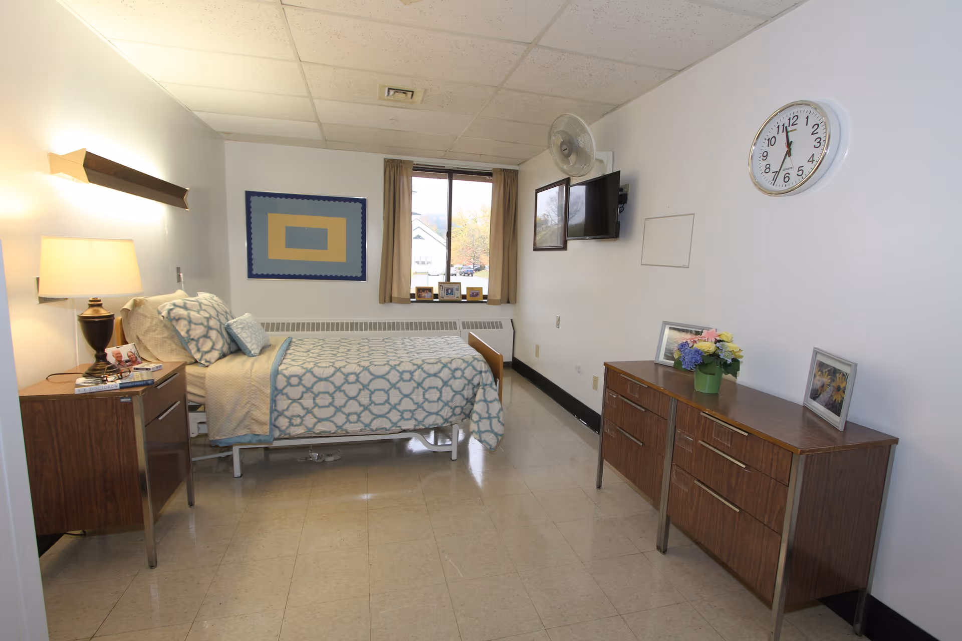 Single-occupancy senior living bedroom with a bed, nightstand, dresser, wall-mounted TV, clock and a window.