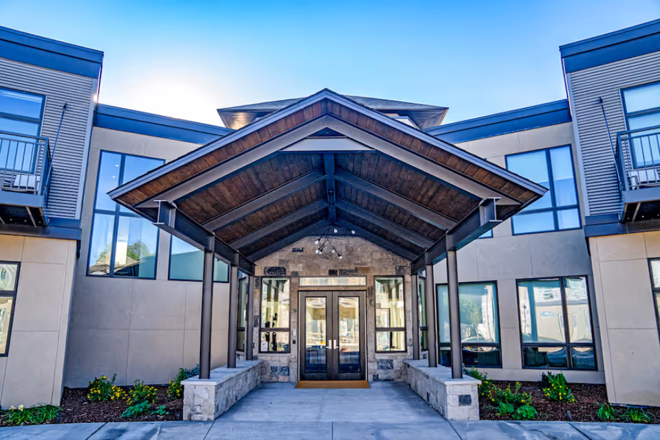 Entrance of Sopris Lodge at Carbondale Senior Living featuring a covered walkway with a wooden roof supported by metal pillars, flanked by beige and gray building walls with large windows and small landscaped garden beds.
