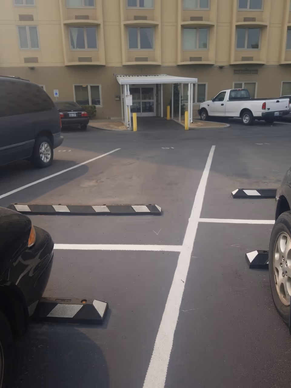 Parking lot with painted spaces leading to the covered entrance of a beige multi-story building.